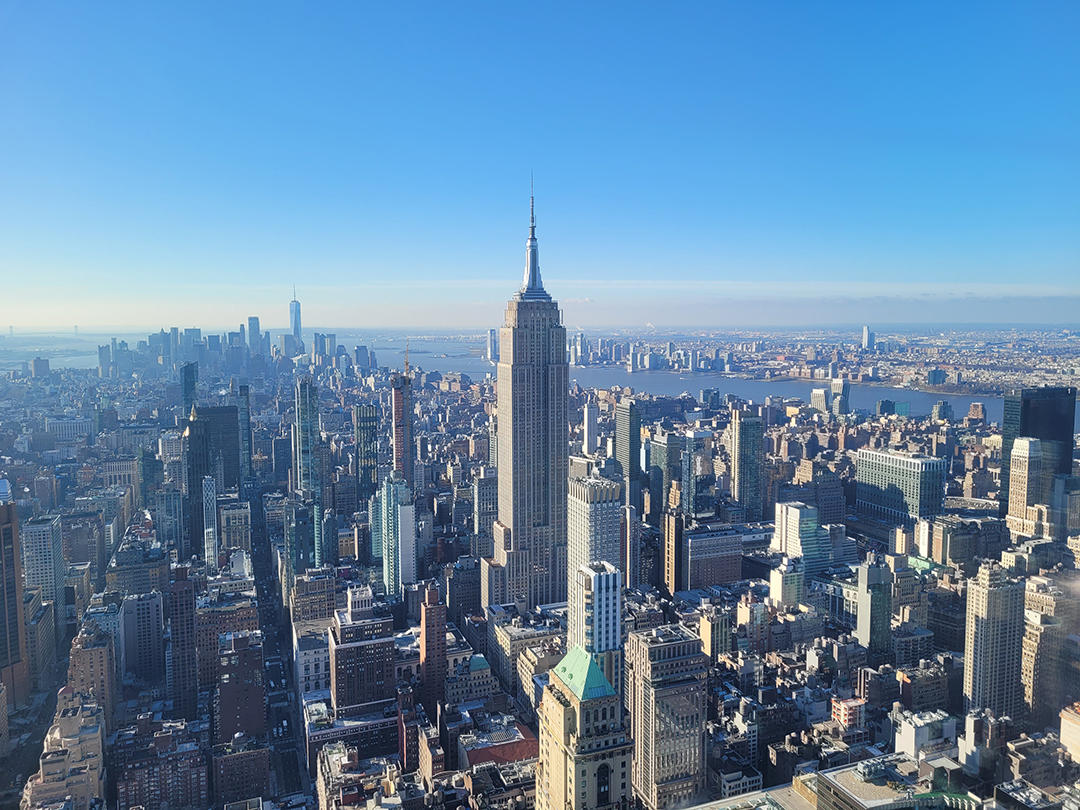 Razgled z vrha stolpnice Empire State Building na Manhattan, kjer mogočna panorama razkriva gosto pozidano srce New Yorka z nebotičniki in reko Hudson v ozadju.