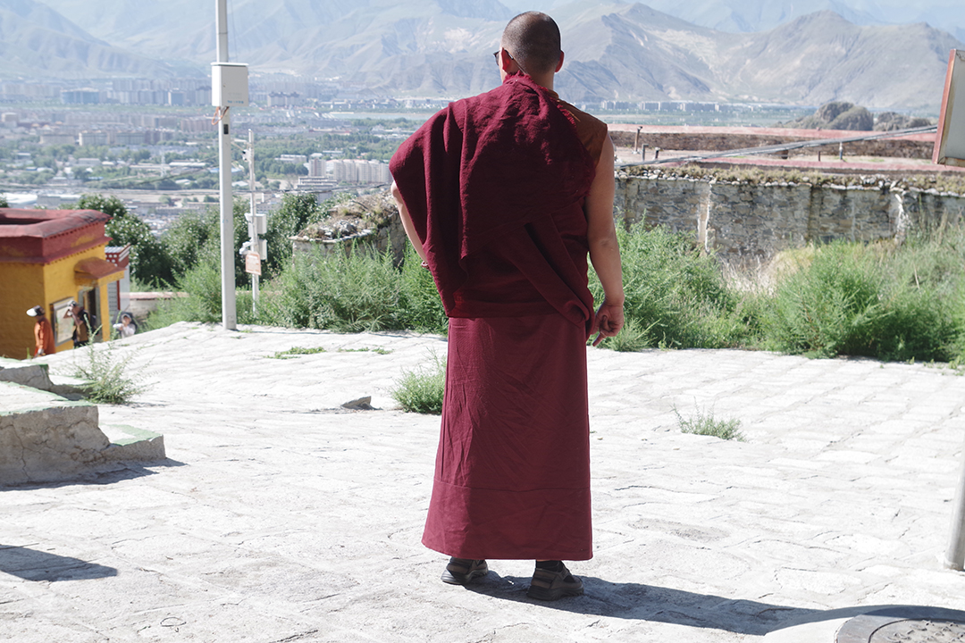 Buddhist monk in traditional red robes overlooking Lhasa and the mountainous Tibetan landscape.